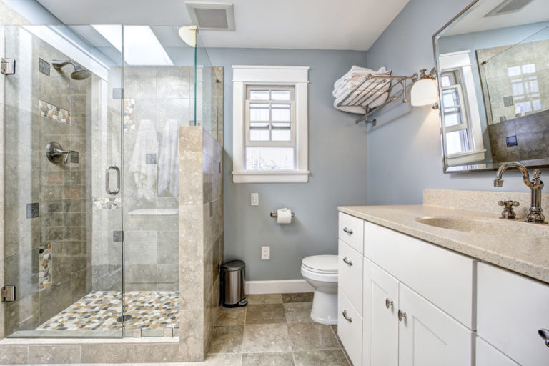 bathroom with glass-enclosed shower on left with skylight above, light gray tile floors, white vanity along right side with tan countertop, light blue/gray walls, and small window open on far wall.