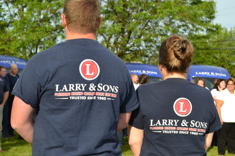 The backs of two people wearing Larry & Sons t-shirts, standing outside watching a community event with trees and other people in background.