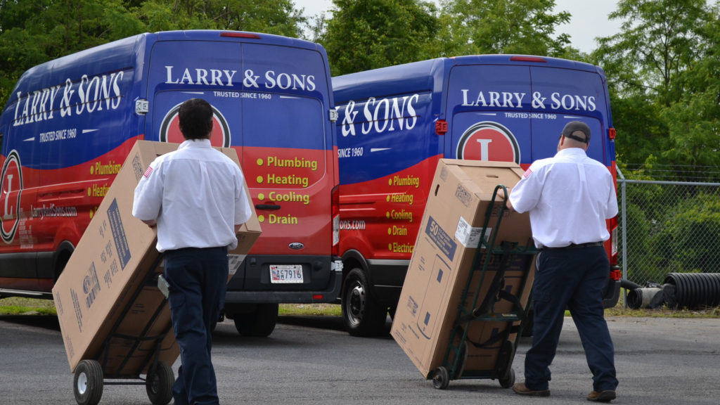 Two Larry & Sons technicians wheeling boxed units out to two service vans parked side by side, with green trees in background.