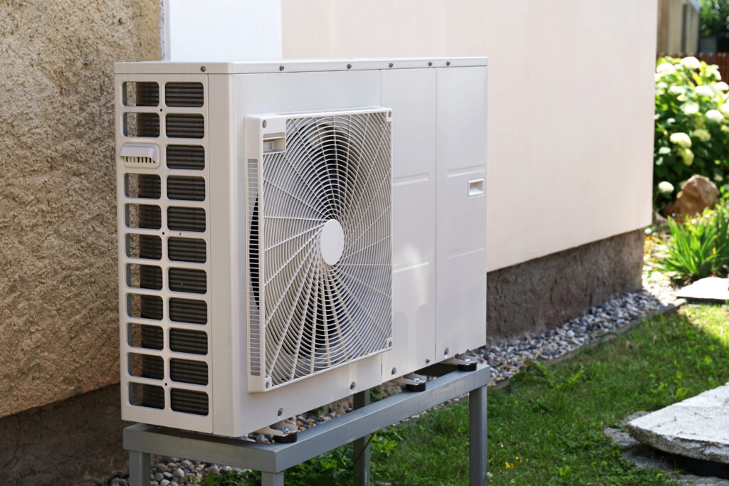 Angled view of an outdoor heat pump unit near the exterior wall of a white stucco house.